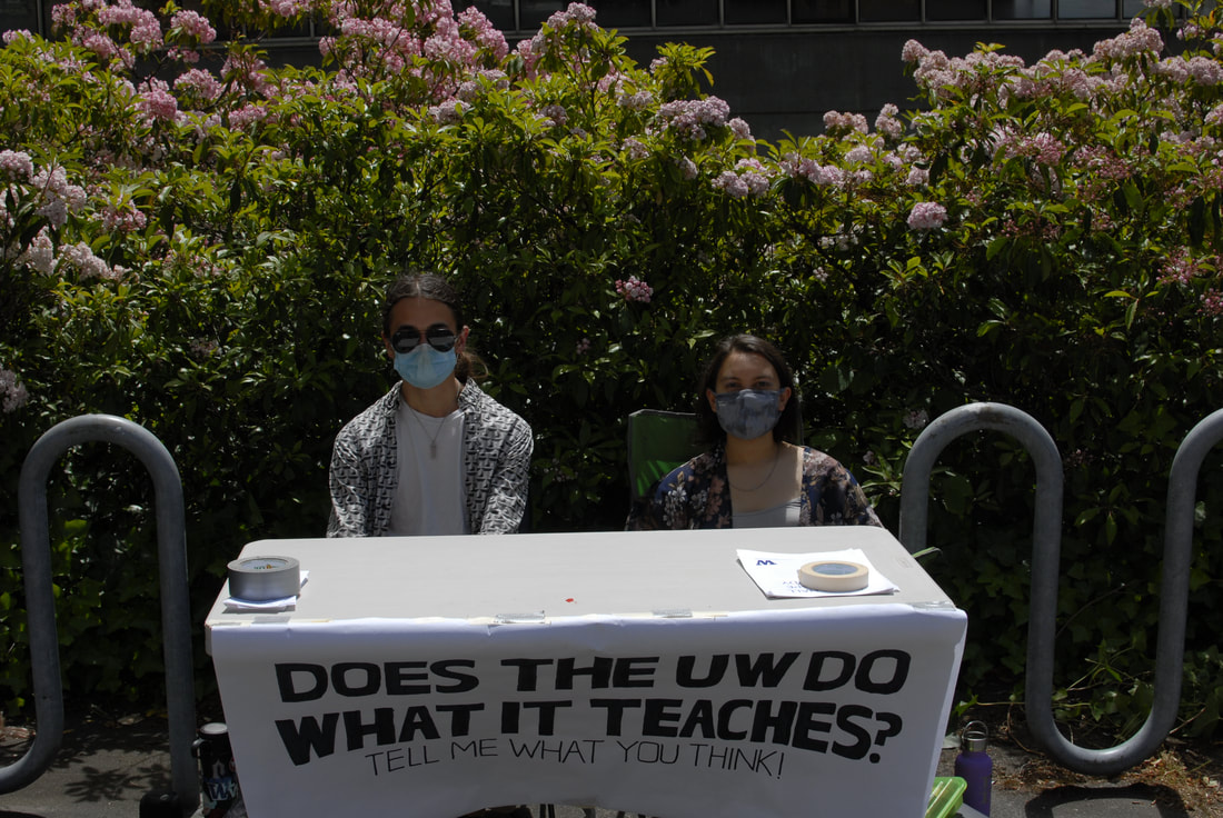 A picture of me sitting behind a table with a sign draped over the edge that reads 'Does the UW do what it teaches?' with a friend who helped with the sign. Pamphlets held down by tape are visible. Bike racks flank us behind us, and calico bushes grow several feet above us furhter back. The concrete facade of Gould Hall and a few windows are visible beyond.