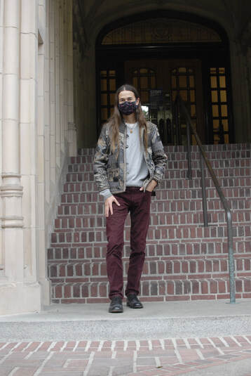 Miguel with a staircase behind them leading up to wood and glass double doors and a detailed off-white stone material on their left. Miguel is wearing a detailed brown and black jacket, a grey sweater, a copper-colored necklace, maroon corduroy pants, and a black and red mask. 