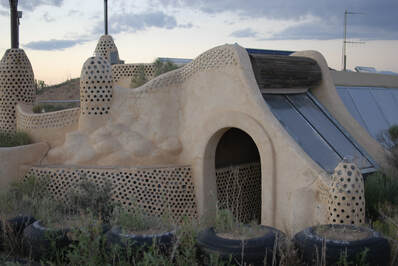 A retaining wall of tires lies in front of a tan structure with many curving forms. Some of these forms meld into cones, and many parts, especially the tops of walls, are decorated with bottles that shine in the light of sunset. A doorway that leads to the building's entrance is visible, and a line of upright windows trails off to the right of the image.